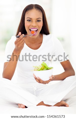 healthy african woman sitting on bed and eating salad