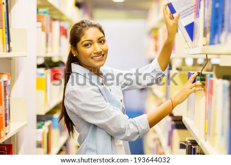happy female indian college student pulling a book off shelf in library