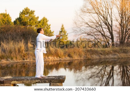 casual young man standing on pier and fishing by the pond