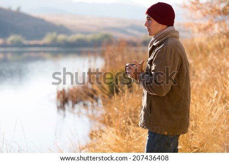 thoughtful man standing by lake and drinking coffee in autumn