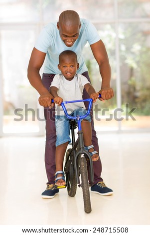 happy african man helping his son to ride a bike indoors
