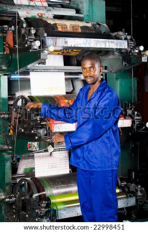 african male machine operator standing next to an old printing press