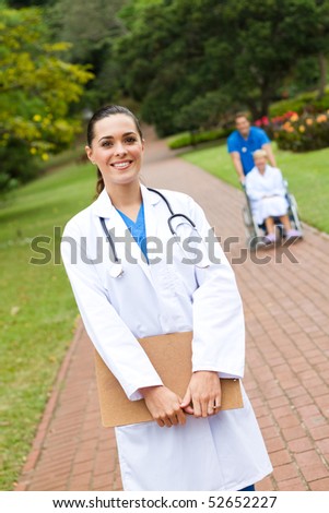 beautiful female doctor portrait outdoors, background is her colleague pushing patient in wheelchair