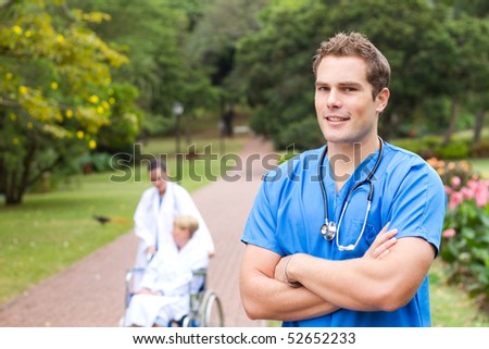 friendly young male doctor portrait outdoors, background is his colleague pushing recovery patient in wheelchair