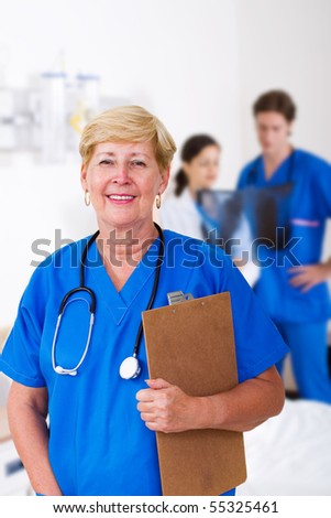 senior nurse with clipboard in hospital, background is her young colleagues