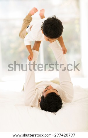playful indian father lying on the bed and lifting up his little son at home