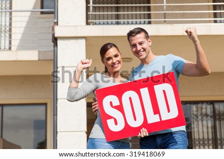 cheerful young couple holding sold sign in front of their old house