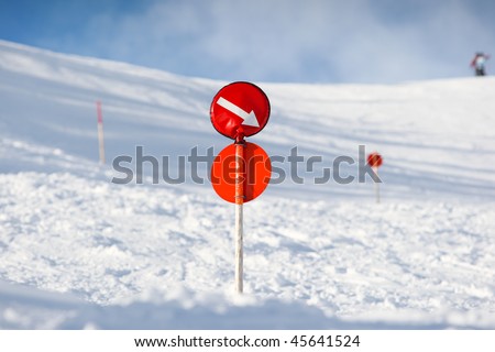 Red sign on a post marking the edge of skiing slope with an arrow showing the way during winter time. More signs and skiers in the blurred background