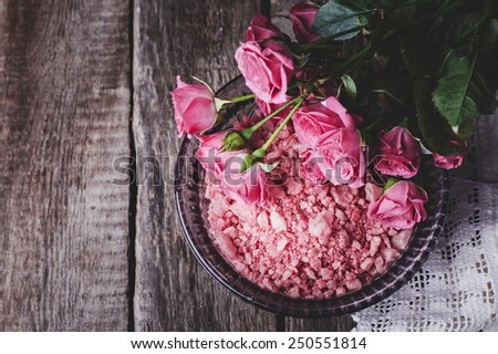 Spa concept with pink sea salt in glass bowl, roses and napkin on vintage wooden background. Selective focus on salt. Toned image