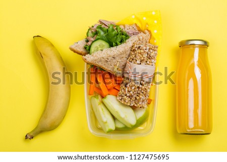 Kids lunch box with sandwich, vegetable sticks, granola bar, banana and juice on yellow background. Back to school concept