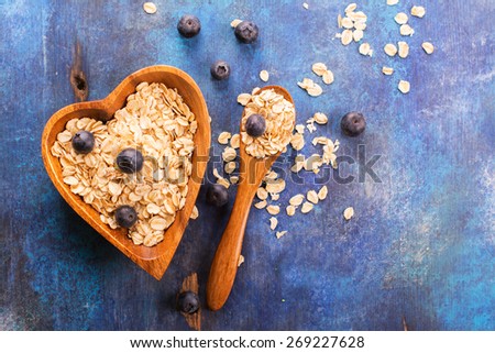 Raw oat flakes with fresh blueberry in heart shaped wooden bowl on blue rustic background. Selective focus