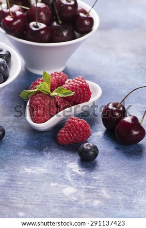 Sweet organic berries: raspberry, blueberry and cherry in white bowls on blue grunge background. Selective focus