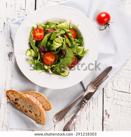 Fresh salad with cherry tomatoes, arugula, cucumber and cheese cubes in white plate on white wooden background. Top view. Square image