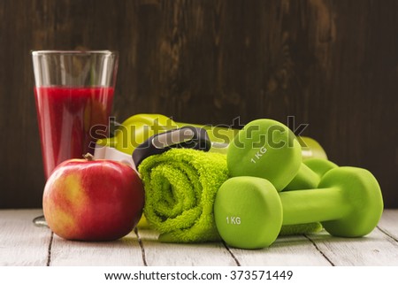 Fitness or diet concept: dumbbells, fresh red smoothie, apple, towel and activity tracker over wooden background. Toned image. Selective focus