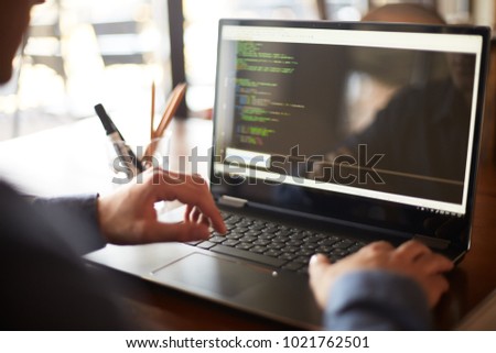 Back view over the shoulder shot of developer programmer with laptop. Program code and script data on the screen. Young freelancer in glasses working on project in cafe.