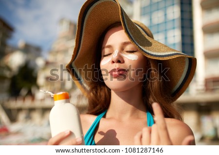 Suntan lotion. Woman applying sunscreen solar cream on face. Beautiful happy cute girl puts suntan cream from plastic container bottle on her nose and cheeks. Female in straw hat and bikini on beach.