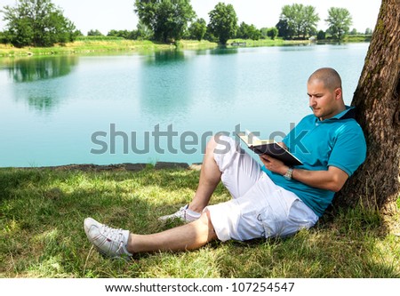 man is sitting outside in a park with lake and reading a book