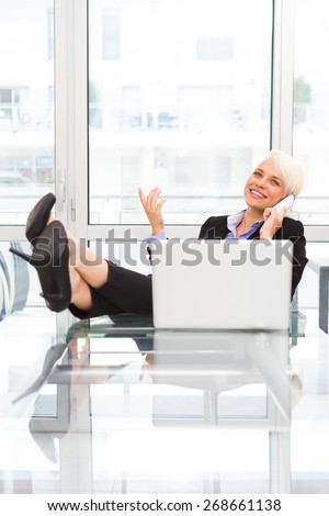 Blonde businesswoman is phoning in the office with legs on the desk