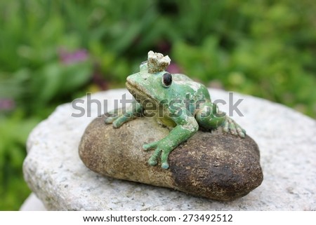 Waiting weathered stone frog statue waiting on a stone to be kissed in the garden, seen from birds eyes view looking up. Character from a Russian fairytale. Black eyes. Blurry green background.