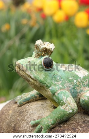 Waiting weathered stone frog statue waiting on a stone to be kissed in the garden, seen from front view. Character from a Russian fairytale. Black eyes. Blurry tulip flower garden background, green.