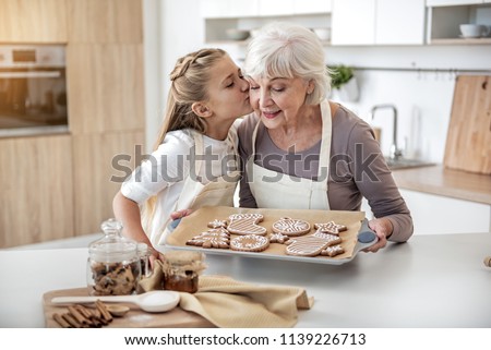 Thank you, granny. Cute girl is kissing cheek of her grandmother with love. Senior woman is holding pray with self-baked cookies and smiling