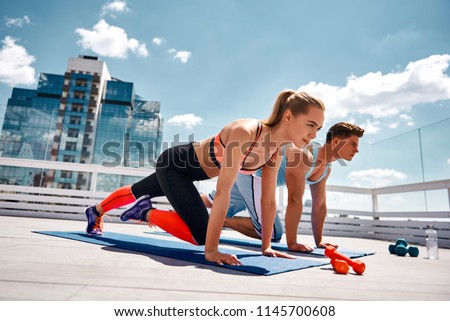 Strong man and woman are doing climber exercise while staying in plank position. They are training together on sunny roof among city center. Enjoying fitness with partner on sunny day concept