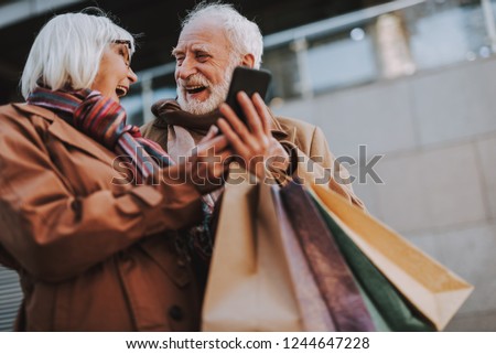 Waist up portrait of cheerful old lady holding cellphone while husband with shopping bags looking at her and laughing
