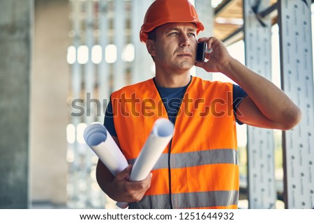 Phone talk. Thoughtful attentive builder in orange uniform standing with the drawings and having a phone talk