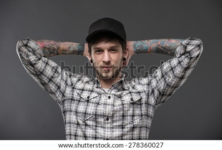 young guy with tattoos and piercings in the ears, holding his hands behind his head and looks directly into the camera. Dressed in a plaid shirt and a black baseball cap