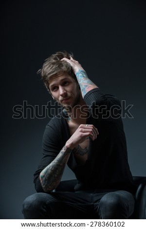 guy with messy hair sitting on a chair. With tattoos on both arms and piercings in the ear, on a black background