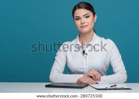 Waist up portrait of pretty woman reporter with brown hair, who is telling news and looking seriously at the camera, sitting at the table on which there is notebook folder and pen, isolated on a blue