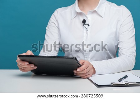 Close up portrait of hands of elegant woman reporter holding the notebook and sitting at the table. There are also pen and folder lying on the table, isolated on a blue background.
