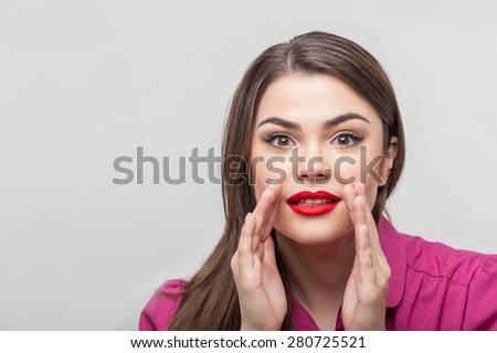 Waist up portrait of beautiful young woman reporter with brown hair and bright red lips, who is very surprised about something while raising up her hands near her mouth and looking at the camera.