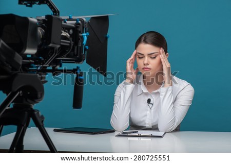 Waist up portrait of elegant woman TV reporter, who is having a headache and keeping her hands on the temples while sitting at the table and her eyes are closed, the camera is visible in the foreground.