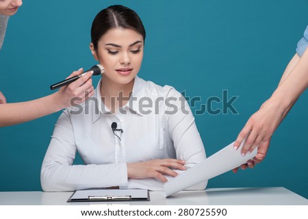 Waist up portrait of beautiful woman reporter with brown hair, who is sitting at the table while looking at the documents, which someone holds in their arms and another woman is doing make up for the