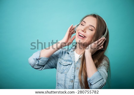 Waist up portrait of beautiful Hipster girl, who is listening to music with the help of headphones and smiling while touching them by both hands and singing a song with her eyes closed.