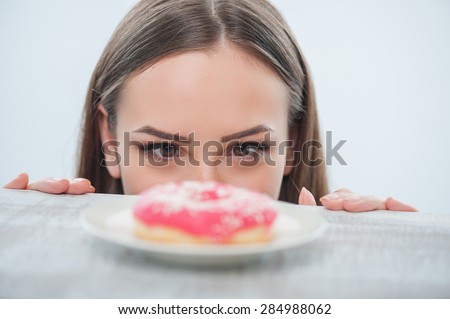 Beautiful girl is looking at unhealthy donut with appetite. It is situated on a table. Isolated on a white background