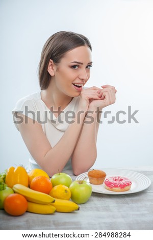 Beautiful girl wants to eat fruits and donuts. She is looking at the camera passionate and smiling. Isolated on a white background
