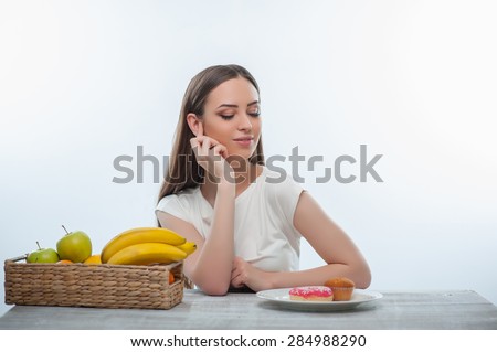 Beautiful girl is looking at unhealthy donuts and can not resist the temptation. She is sitting at the table. Isolated on a white background