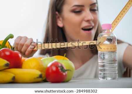 Pretty woman is measuring bottle of water with tape-line seriously. Near her fruits and vegetables are situated. Isolated on a white background