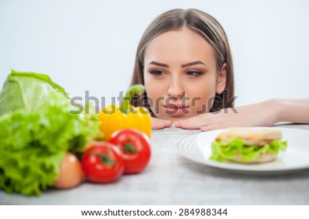 Pretty woman is making decision to eat vegetables. She is looking at it and does not pay her attention on hamburger. Isolated on a white background