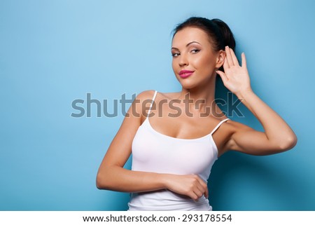 Attractive woman is raising her palm to her ear to overhear some interesting news. She is smiling wily and looking aside. Isolated on blue background and there is copy space in left side