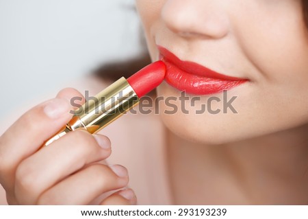 Close up portrait of attractive lips of beautiful woman. She is rouging her lips with red lipstick. The lady is gently smiling. Isolated on grey background