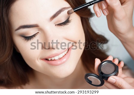 Attractive woman closes her eyes and smiling. The female hands are holding the eye shadow and brush. Female hand makes-up her eyes. Isolated on grey background