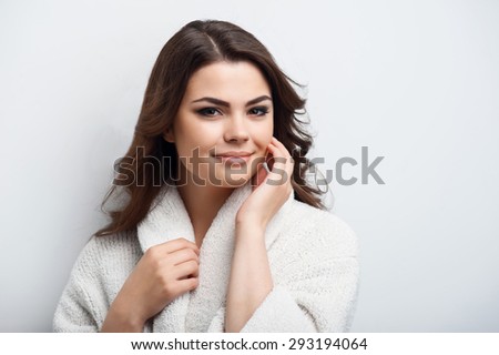 Waist up portrait of beautiful woman in bathrobe. She is looking mysteriously at the camera and slightly smiling. The lady is touching her face. Isolated on a grey background and there is copy space