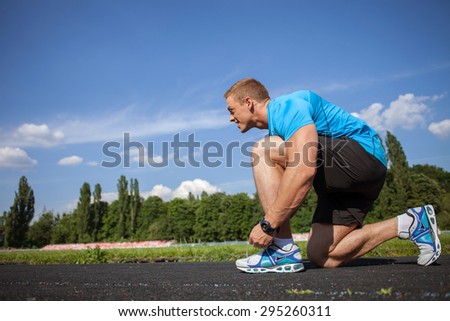 Attractive healthy man ties the laces in stadium. He takes a seat and bents his knee in profile. The sportsman is looking forward with desire. There is copy space in left side