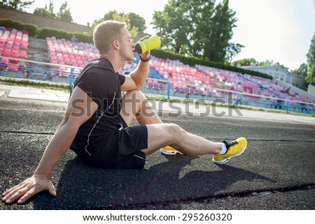 Cheerful athlete is sitting on road in stadium. He is drinking water from bottle. His eyes are closed with pleasure
