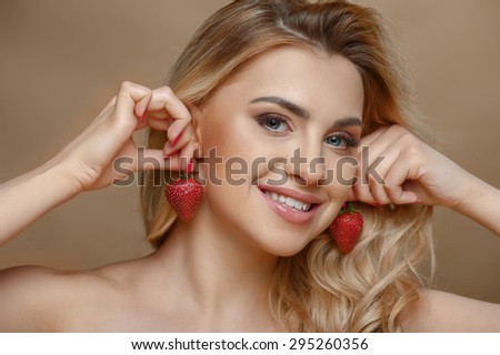 Cheerful young woman is touching two strawberries to her ears. She is smiling and making fun. Isolated on brown background