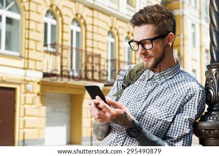 Cheerful hipster guy is holding mobile phone and looking at it with interest. He is gently smiling in city