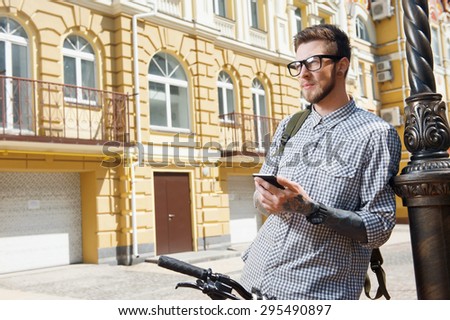 Handsome young hipster guy is standing near bicycle. He is holding mobile phone and looking seriously and thoughtfully aside. He has backpack on his shoulder